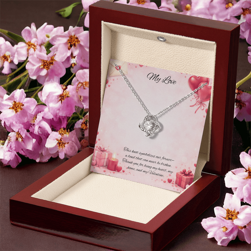 Silver love knot necklace with a sparkling center stone displayed in a red gift box on a soft Valentine-themed background with hearts and romantic message card.