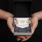 Close-up of hands holding an open red gift box featuring a personalized silver name necklace with heart, set against a message card that reads "Graduation 2025" and includes an inspiring graduation quote, with gold and white balloons in the background.