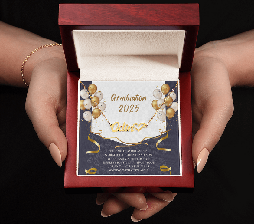 Close-up of hands holding an open red gift box featuring a personalized gold name necklace with heart, set against a message card that reads "Graduation 2025" and includes an inspiring graduation quote, with gold and white balloons in the background.