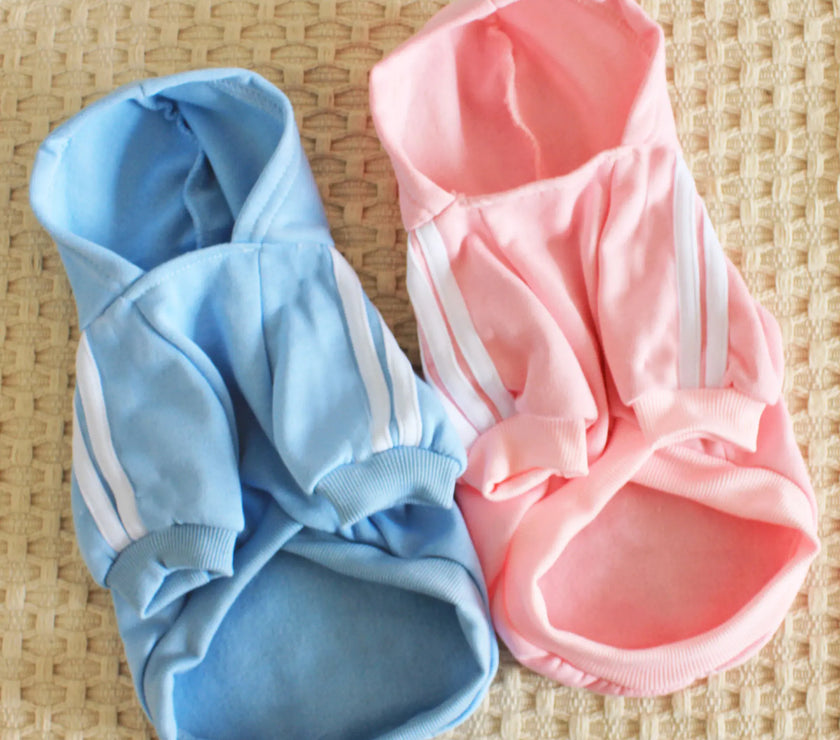 Two dog hoodies, one blue and one pink, on a woven surface.